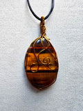 Colgante alargado de ojo de tigre con bandas doradas y marrones, engarce de cobre en espirales y cordón negro