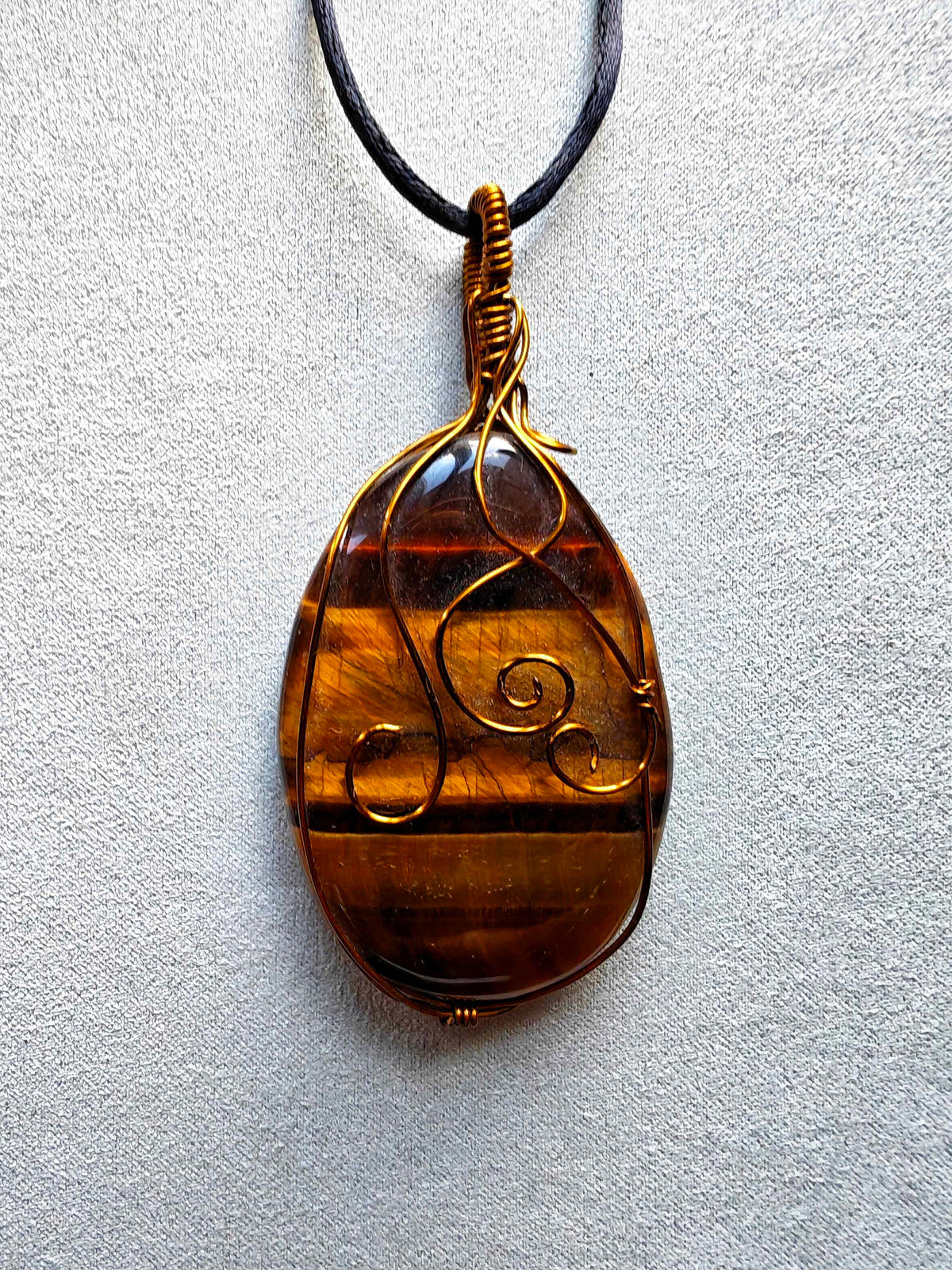 Colgante alargado de ojo de tigre con bandas doradas y marrones, engarce de cobre en espirales y cordón negro
